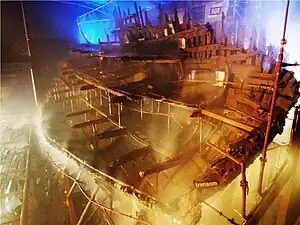 A diagonal section of a wooden ship seen from the stern inside a moodily lit building while it is being sprayed with water from a sprinkler system