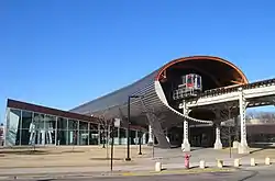  large steel tube encircling elevated train track with a train on it, over low building with large glass windows