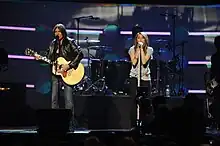 A long shot of a female performer, with long brown hair, singing into a microphone. Both of her hands rest on a microphone stand decorated with jewels. To her right is a male musician wearing a leather jacket, playing an acoustic guitar and also singing into a microphone.