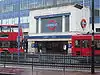 A grey building with a rectangular, dark blue sign reading "MORDEN STATION" in white letters, two red buses in front, and a fence in the foreground