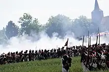 Photo shows a re-enactment of a Napoleonic battle. A French firing line is seen from the rear while a church tower looms to the right.