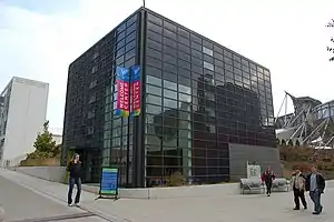 A cube-shaped building covered by a rectangular grid containing some windows, but mostly black glass. Four people are on the surrounding sidewalks and there are three banners reading "Welcome Center".