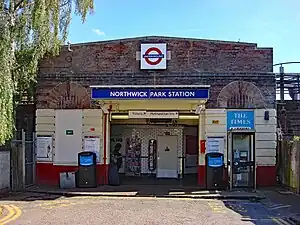 A brown-bricked building with a rectangular, dark blue sign reading "NORTHWICK PARK STATION" in white letters all under a light blue sky