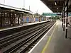 A grey-tiled railway platform bordered by red-bricked walls supporting overhanging roofs from which elongated electrical lights hang