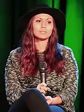 A brunette woman is seated at a chair; she is wearing a hat.