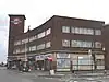 A brown-bricked building with a white sign reading "ZACHARIA FASHIONS WHOLESALERS" in blue letters all under a blue sky with white clouds