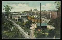 A 1911 postcard showing a quarrying operation in Portland Connecticut with cliffs of brownstone in the background, rail lines for loading and transporting stone, industrial buildings, rail carts, and other parts of the operation.