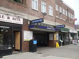 A brown-bricked building with a rectangular, dark blue sign reading "QUEENSBURY STATION" in white letters all under a light blue sky