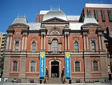 A 3 story, brick and red stone, second empire style building with a slate roof. The building is symmetrical with the entrance center and a blue and yellow banner on either side.