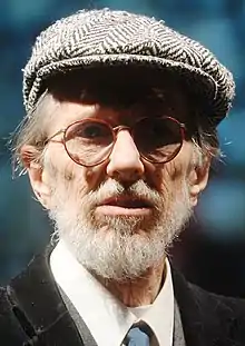 An elderly man with a white beard, round glasses, a beret-like hat, a dark vest, and a necktie.  He faces down right, looking into an open book.