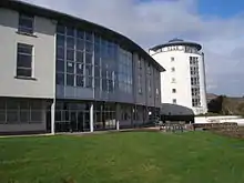 A modern 3 story building with a prominent frontage of numerous windows and constructed from a white material curves gently away from a green lawn in the foreground. In the background there is a tall white tower of a similar construction.