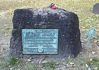 A rectangular, rough-hewn block of stone on the ground. There is a walkway in the foreground, grass and trees in the background. A weathered plaque on the stone reads: "Samuel Adams, Signer of the Declaration of Independence, Governor of this Commonwealth, A Leader of Men and an ardent Patriot."