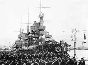 A black and white picture of a ship at anchor with uniformed men to the ship's left side.