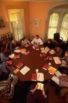  eleven people sitting around octagonal wood table with red books in front of them seen from above