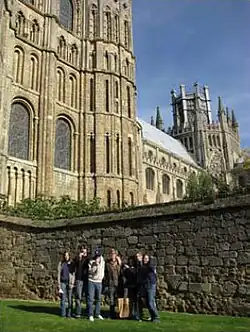  seven young people standing on grass in front of stone wall with Gothic building behind them