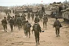 Men in desert-brown fatigues walk towards rows of tanks. All of them have backpacks, and some have firearms.