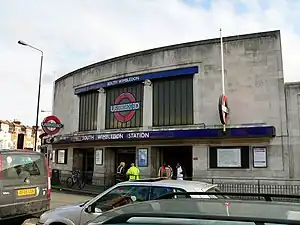 A grey building with a rectangular, dark blue sign reading "SOUTH WIMBLEDON STATION" in white letters all under a light blue sky