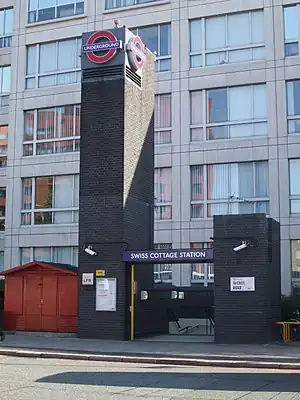 Two black-bricked pillars on a sidewalk with a rectangular, dark blue sign connecting them reading "SWISS COTTAGE STATION" in white letters