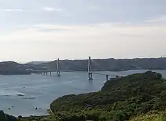 白色的雙塔斜張橋跨越藍色的日比水道,海岸線崎嶇而青翠。陽光和煦,幾隻小艇在泛起細碎波紋的海面上游弋。