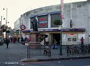 A grey-bricked building with a rectangular, dark blue sign reading "TOOTING BROADWAY STATION" in white letters all under a grey sky