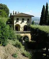 A yellow stucco building with a red clay roof and several arched doors and windows, surrounded by green shrubs.