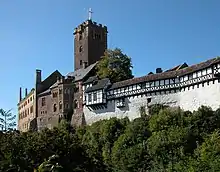 A castle perched along the edge of a wooded hill. The castle has grown in several stages and consists of sections in dark stone, lighter stone, white plaster and half-timber.