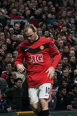 A man with short, thinning hair, wearing a red shirt with a black collar and a shallow black chevron on the chest, and white shorts.