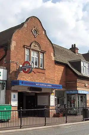 A brown-bricked building with a rectangular, dark blue sign reading "WEST HAMPSTEAD STATION" in white letters all under a blue sky