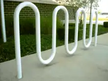 A white-painted round metal tube bent into a double M shape and set in concrete in front of a grassy verge and a brick wall.