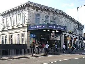 A white-bricked building with a rectangular, dark blue sign reading "WILLESDEN GREEN STATION" in white letters all under a light blue sky