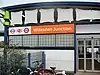 A white-panelled building with a rectangular, orange sign reading "Willesdon Junction" in white letters all under a light blue sky