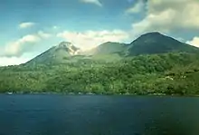 A photograph depicting a blue sky at the top, a grey mountain range in the middle, white clouds in front of the mountain range, and a rocky terrain at the bottom.